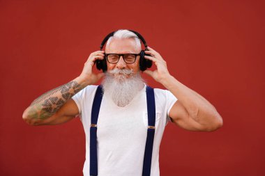 Beyaz sakallı ve bıyıklı yaşlı adam kulaklıklı favori müziği dinliyor. İnsanlar mutlu bir yaşam tarzı ve teknoloji konsepti. Kırmızı arka plan. Modaya uygun olgun erkekler.