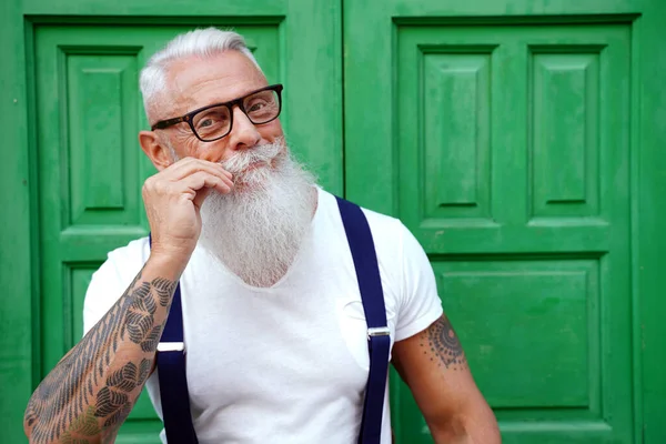Handsome senior man with white beard and mustache wearing fashionable suspenders and eyeglasses, looking at the camera, posing on the green door.