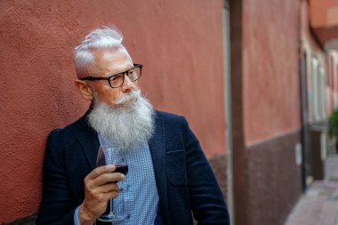 Beyaz sakallı, bıyıklı, elinde kırmızı şarap kadehi olan yakışıklı, modaya uygun bir son sınıf öğrencisi. Açık hava fotoğrafı. Zarif emekli poz veriyor.