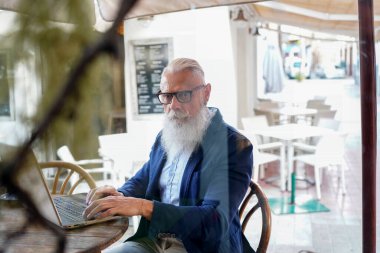 Şık sakallı kıdemli işadamı iş yerinde bilgisayarını kullanıp kameraya bakıyor. İş yerinde yakışıklı bir işadamı