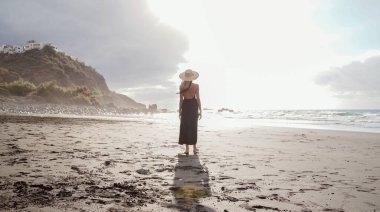 Gün batımında boş kumsalda yürüyen, yaz şapkası takan, okyanusa bakan güzel romantik bir kadın. Tatil adası titreşimleri.