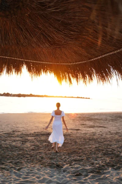 日出时穿着白色淡紫色连衣裙在海滩上奔跑的女子的背影 真正的人复古风格 — 图库照片