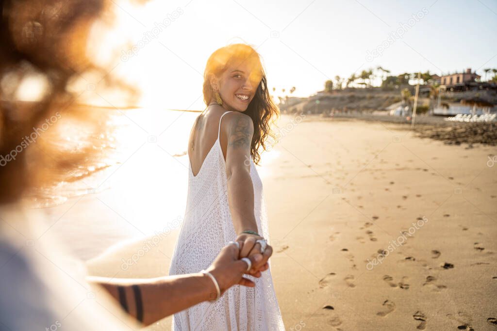 Lovely smiling beautiful woman holding hand of her boyfriend, looking at him, walking on the beach during sunset. Summer island vibes. Love is in the air. Real people lifestyle.
