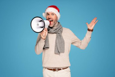 Noel Baba 'nın kırmızı şapkasını takan yakışıklı mutlu adam megafonla iyi haberler bağırıyor. Mavi Pastel stüdyo arka planı. Noel zamanı. Xmas satışları.