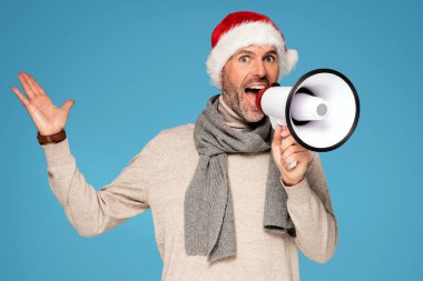 Noel Baba 'nın kırmızı şapkasını takan yakışıklı mutlu adam megafonla iyi haberler bağırıyor. Mavi Pastel stüdyo arka planı. Noel zamanı. Xmas satışları.