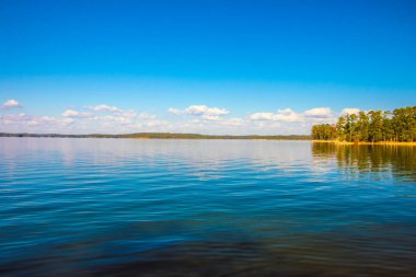Mavi gökyüzü ve suda yansıyan bulutlarla güzel bir göl manzarası.