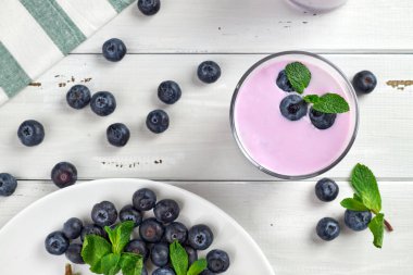 Beyaz ahşap bir masada taze yaban mersini ve nane yapraklarıyla servis edilen yaban mersinli yoğurt. Sağlıklı kahvaltı. Üst görünüm.