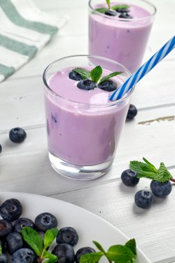 Beyaz ahşap bir masada taze yaban mersini ve nane yapraklarıyla servis edilen iki bardak yabanmersinli yoğurt. Sağlıklı kahvaltı..