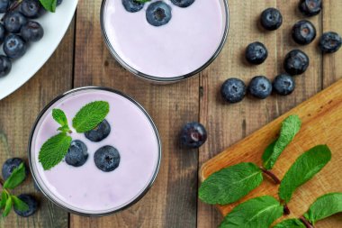 Taze meyve ve nane yapraklı yaban mersinli yoğurt. İki bardak yoğurt ve ahşap bir masada bir sürü yaban mersini. Sağlıklı kahvaltı..