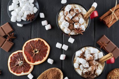 Two red cups of hot chocolate with marshmallow, anise and cinnamon sprinkled with cocoa powder on a wooden table with slices of dried grapefruit and chocolate pieces. Winter hot drink top view.
