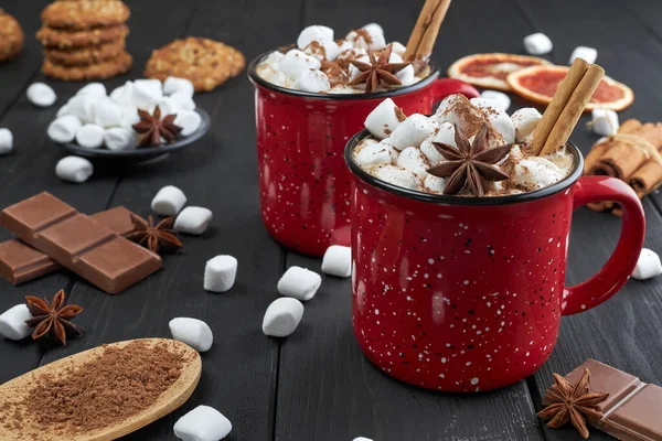 Two red cups of hot chocolate with marshmallow, anise and cinnamon sprinkled with cocoa powder on a wooden table with slices of dried grapefruit and chocolate pieces. Winter hot drink