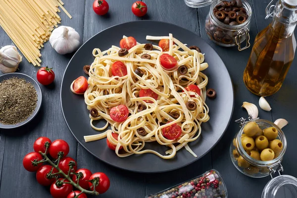 Delicious Italian pasta tagliatelle with cherry tomatoes and basil on a plate on a wooden table. Ingredients for making pasta: olives, tomatoes, garlic, olive oil and various types of dried peppers.