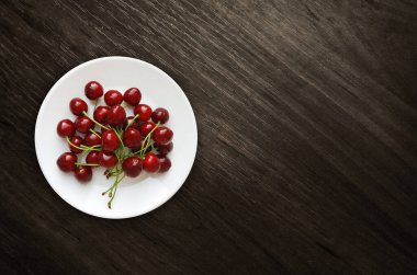 Kahverengi ahşap zemin üzerinde beyaz yuvarlak bir tabakta kırmızı kirazlar ve fotokopi alanı. Üst görünüm