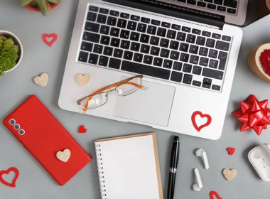 Kırmızı cep telefonu, not defteri, gözlükler ve sevgililer günü dekorasyonları. Gri masadaki dizüstü bilgisayarın yanında.