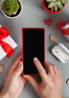Hediye kutularının yanındaki akıllı telefonu kullanan eller ve gri masanın üstündeki kalpler.