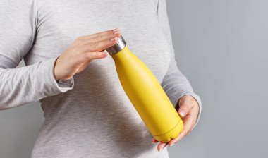 Gri tişörtlü, elinde sarı yalıtımlı şişeyle gri arka planda bir kadın.