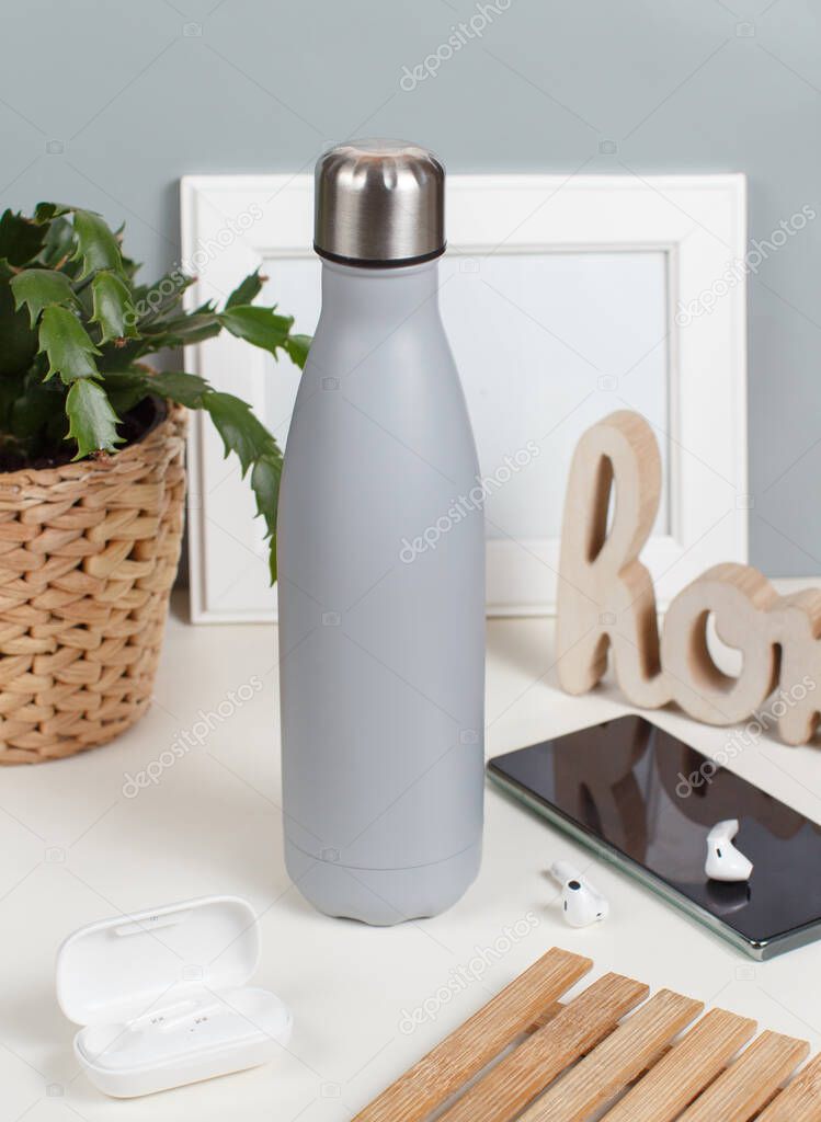 Grey insulated bottle on white desk surrounded by modern gadgets and plant front view