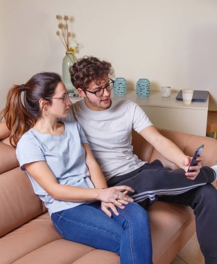 Genç kadınlar ve erkekler evdeki bej kanepede otururken cep telefonlarıyla video görüşmesi yapıyorlar. Adamların elinde akıllı telefon var.. 