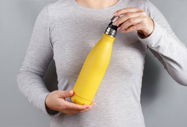 Gri tişörtlü, elinde sarı yalıtımlı şişeyle gri arka planda bir kadın.