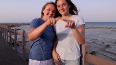 Anne ve kızı denizin kenarında duruyor ve kalplerini gösteriyor.