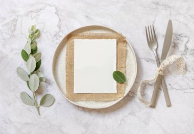 Beyaz tabakta, çatal ve bıçakla okaliptüs dalları ve klasik kurdelelerle dolu mermer masanın üzerinde duran boş bir kağıt kart. Düğün davetiyesi maketi