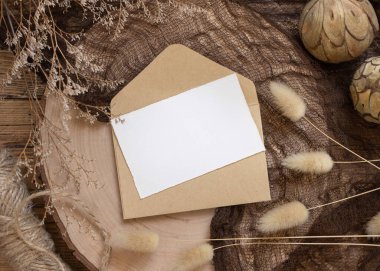 Zarfın üzerinde boş bir kağıt kart ve etrafında kurumuş bitkiler olan ahşap bir masa. Davetiye şablonlu Boho model sahnesi