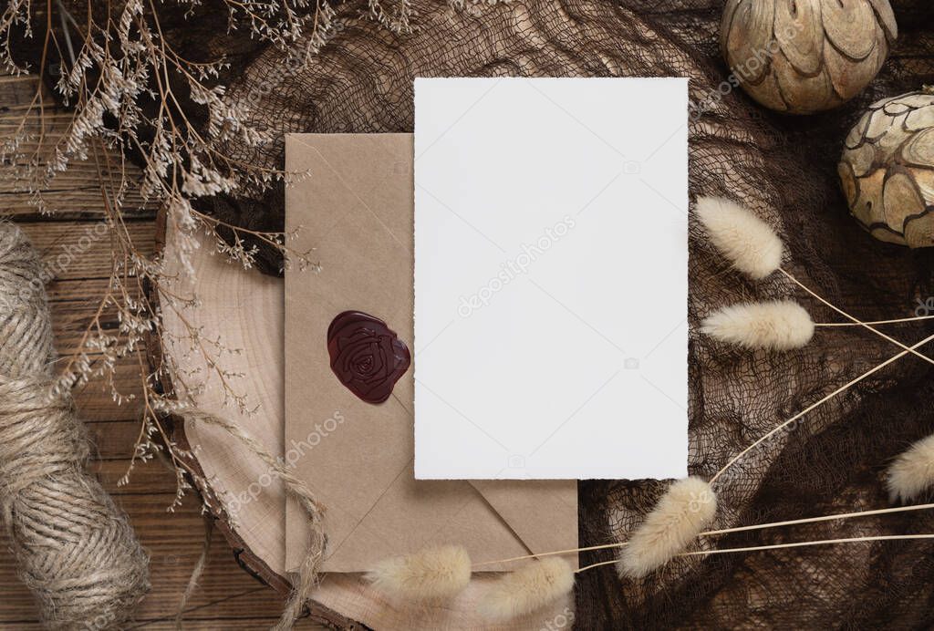 Blank paper card and envelope on a dark wooden table with dried plants around, top view. Boho mock-up scene with invitation card template