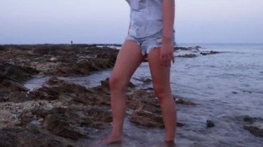 Açık mavi tişört ve kot şort giyen genç kız gün batımında deniz kenarında bir uçurumda yalınayak yürüyor.