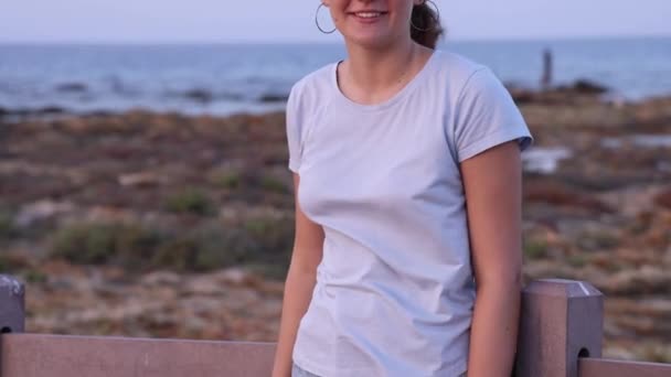 夕日と笑顔で海のそばの木製の歩道に立って10代の女の子 薄い青のTシャツ わらの帽子と財布を身に着けている若い女性 夏の旅行のコンセプト — ストック動画