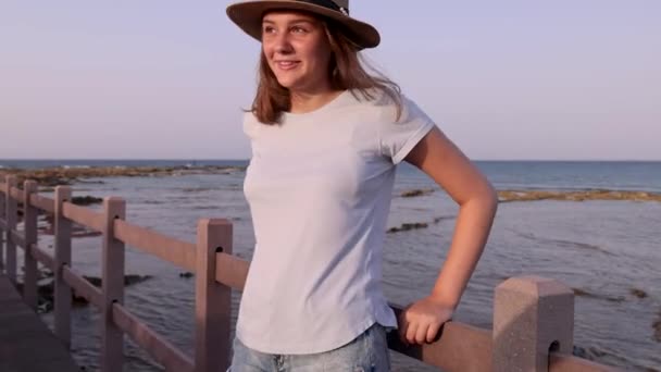 日落时 站在海边的木制人行道上的少女突然出现了 穿着浅蓝色T恤 草帽和钱包的年轻女子 暑期旅行概念 — 图库视频影像