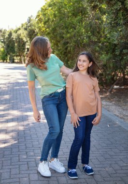 Anne ve kızı parkta yürüyor. Orta yaşlı bir kadın ve çocuk el ele tutuşuyor, göz teması kuruyor ve yeşil ve bej tişörtler giyiyor.