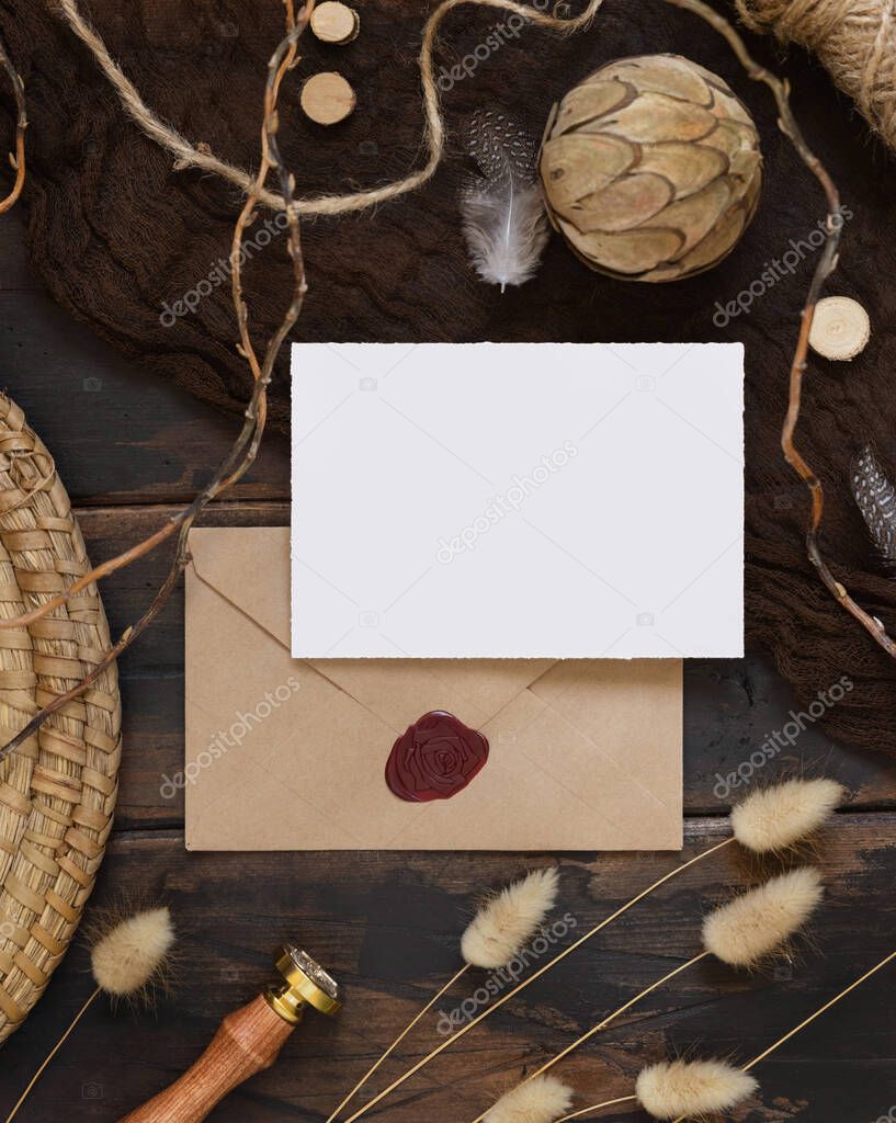 Blank paper card and envelope on a dark wooden table with dried plants around, top view. Boho mock-up scene with invitation card template