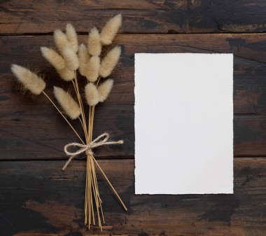 Kahverengi ahşap masanın üzerinde boş beyaz kağıt kart, yanında da kurutulmuş çiçek buketi, üst manzara. Boho davetiye kartı modeli