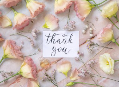 Üzerinde pembe çiçekler, küpeler, taç yaprakları ve misket masasının üzerindeki parfüm kaplamalarıyla çevrili el yazısıyla yazılmış teşekkür kartı. Romantik konsept