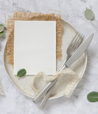 Beyaz tabakta boş kağıt kart, çatal ve bıçakla mermer masanın üzerinde okaliptüs dalları ve klasik kurdeleler, üst manzara. Kart modeli