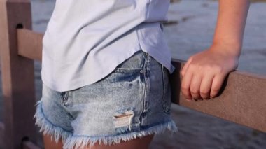 Gün batımında denizin kenarında duran genç kadına yaklaş. Açık mavi tişört ve kot şort giyen genç bir kadın. Yüzsüz atış. Yaz yolculuğu konsepti