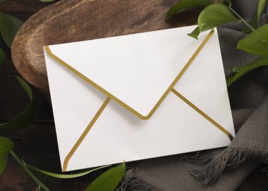 Yeşil yapraklar ve gri kumaşların yanında kahverengi ahşap bir tepside boş altın kenarlı bir zarf. Bohem ya da kırsal düğün davetiyesi zarflı Boho sahnesi