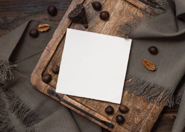 Gri keten kumaş ve doğal dekor elementlerinin yanındaki tahta tepside boş bir kart modeli. Kare kartlı Boho dairesi, bohem ya da doğaya yakın düğün davetiyesi zarfları.