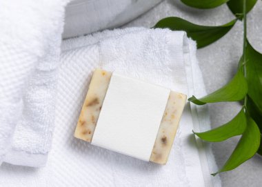 Krem rengi sabun kalıbının ambalajı. Beyaz banyo havlusunun üzerinde boş etiket ve gri taş tezgahın üstünde yeşil yapraklar. Doğal el yapımı bitkisel hijyen ürünü. Tarafsız tuvalet