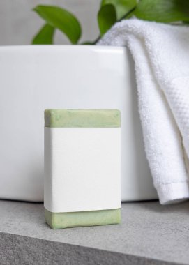 Havza üzerinde boş etiketi olan sabun çubuğu, yeşil yapraklı dal ve gri taş tezgahın üzerinde beyaz banyo havluları kapanıyor, ambalaj modeli. Doğal el yapımı bitkisel hijyen ürünü. Tarafsız tuvalet