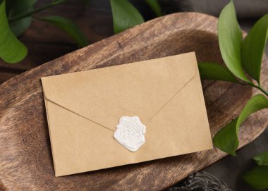 Yeşil yapraklar ve gri kumaşların yanındaki kahverengi ahşap tepside boş mühürlü bir zarf. Kare zarflı boho sahnesi. Bohem ya da kırsal düğün davetiyesi süiti