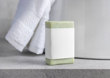 Green soap bar with blank label near white bath towels and basin on grey stone countertop, close up, packaging mockup. Natural handmade hygiene product. Clean neutral bathroom interior