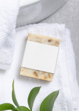 Krem rengi sabun kalıbının ambalajı. Beyaz banyo havlusunun üzerinde boş etiket ve gri taş tezgahın üstünde yeşil yapraklar. Doğal el yapımı bitkisel hijyen ürünü. Tarafsız tuvalet