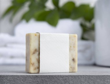 Yeşil yapraklı, beyaz banyo havluları ve gri taş tezgahın üzerinde boş etiketli krem sabun kalıbı kapatılıyor, ambalaj modeli yapılıyor. Doğal el yapımı bitkisel hijyen ürünü. Tarafsız tuvalet