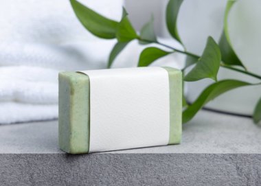 Yeşil etiketli yeşil sabun çubuğu ile yeşil yapraklı beyaz banyo havluları ile gri taş tezgahın üzerinde yakın çekim, ambalaj modeli. Doğal el yapımı bitkisel hijyen ürünü. Tarafsız tuvalet