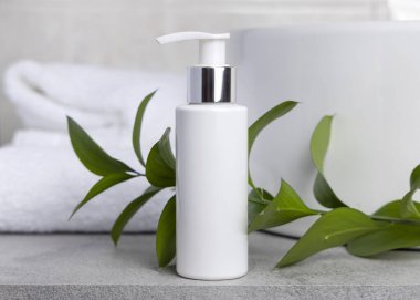 Paket maketi, beyaz boş bir pompa krem şişesi yeşil yapraklı dal, leğen ve beyaz banyo havlularına karşı banyodaki gri taş tezgahın üstünde. Doğal bitkisel kozmetik