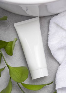 Paketleme modeli, yeşil yapraklı dalların yanında beyaz sıkma krem tüpü, leğen ve beyaz banyo havluları banyo manzaralı gri taş tezgahın üzerinde. Doğal bitkisel kozmetik