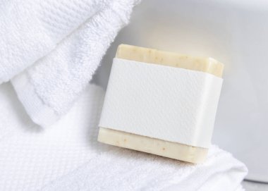 Havlunun yanındaki beyaz banyo havlularının üzerinde boş etiketli krem sabun kalıbı, yakın çekim, ambalaj modeli. Doğal el yapımı hijyen ürünü. Temiz tarafsız banyo, yumuşak ışık.