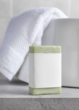 Beyaz banyo havlularının yanında boş etiketli yeşil sabun barı ve gri taştan tezgahın üstünde leğen, yakın plan, ambalaj modeli. Doğal el yapımı hijyen ürünü. Temiz tarafsız banyo içi.