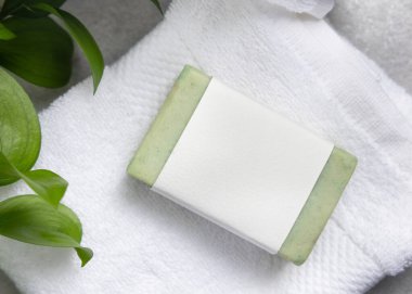 Gri taş tezgahın üzerindeki yeşil yaprakların yanında beyaz banyo havlusunun üzerindeki boş etiketli yeşil sabun kalıbının ambalajı. Doğal el yapımı bitkisel hijyen ürünü. Tarafsız tuvalet